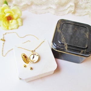 May include: A gold locket necklace with a round, open locket and a delicate chain. The necklace is displayed on a white box with three small gold beads. The necklace is packaged in a black and gold square tin box.