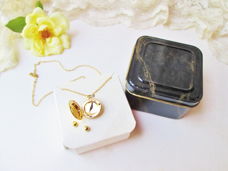 May include: A gold locket necklace with a round, open locket and a delicate chain. The necklace is displayed on a white box with three small gold beads. The necklace is packaged in a black and gold square tin box.