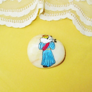 May include: Hand-painted rock art depicting a person in a blue robe and a red sash, holding a staff and a sheep. The person is wearing a yellow crown and has a white beard. The rock is white with brown veins.