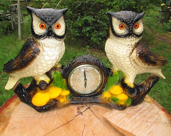 May include: A vintage owl clock featuring two ceramic owls with brown and cream-colored feathers and bright orange eyes. The clock face is round and beige, set between the owls on a brown branch with yellow mushrooms and green foliage.