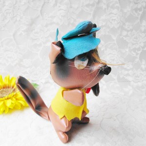 Puede incluir: Un juguete de peluche de ardilla marr&oacute;n y negro que lleva un sombrero azul y una camisa amarilla. La ardilla tiene una cola larga y una nariz negra.