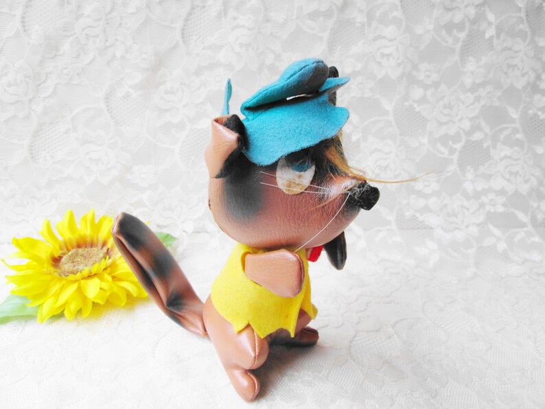 Puede incluir: Un juguete de peluche de ardilla marr&oacute;n y negro que lleva un sombrero azul y una camisa amarilla. La ardilla tiene una cola larga y una nariz negra.