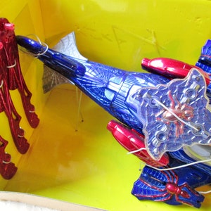 May include: A blue and red toy spider vehicle with a clear plastic spider web design. The vehicle has red wings and a clear plastic spider web design. The vehicle is in a yellow box with red and silver toy spiders.