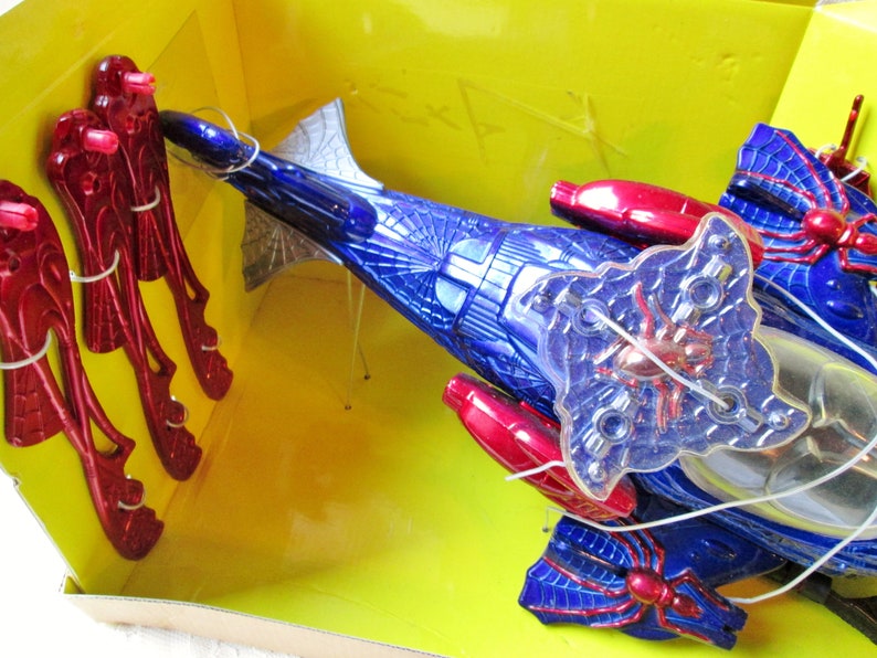 May include: A blue and red toy spider vehicle with a clear plastic spider web design. The vehicle has red wings and a clear plastic spider web design. The vehicle is in a yellow box with red and silver toy spiders.