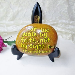 May include: A painted rock with the bible verse "For we walk by faith, not by sight: Paul 2 Corinthians 5:7" in yellow lettering on a brown background. The rock is on a black stand.