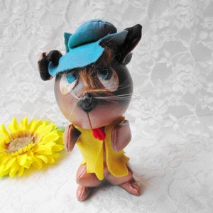 Puede incluir: Un gato de peluche marr&oacute;n que lleva un sombrero azul y una chaqueta amarilla. El gato tiene una corbata de lazo roja y bigotes blancos.