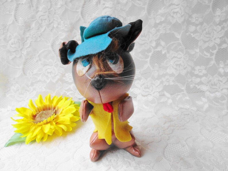 Puede incluir: Un gato de peluche marr&oacute;n que lleva un sombrero azul y una chaqueta amarilla. El gato tiene una corbata de lazo roja y bigotes blancos.