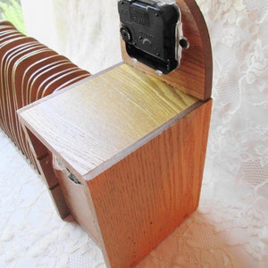 May include: A wooden clock with a clock face and a wooden case. The clock is made of light brown wood and has a dark brown clock face. The clock is sitting on a white surface.