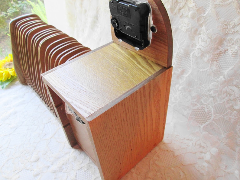 May include: A wooden clock with a clock face and a wooden case. The clock is made of light brown wood and has a dark brown clock face. The clock is sitting on a white surface.