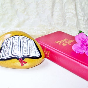 May include: A painted rock with a closed book design. The rock is yellow with a black outline and red ribbon. A red book with gold lettering that reads "Hymns of Faith" is in the background.