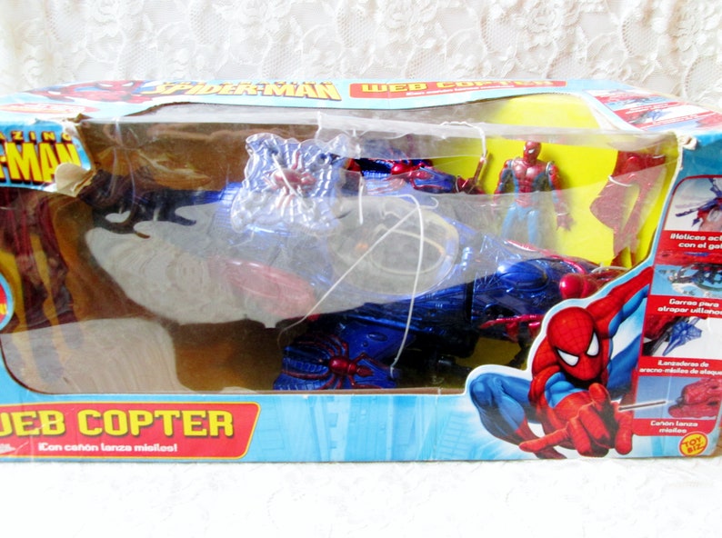 May include: A blue and red toy helicopter with a spider web design. The helicopter has a red and blue spider figure on the front and a red and blue spider figure on the back. The helicopter is in a plastic package with a red and blue Spider-Man figure on the front. The package says "Web Copter" and "Con ca&ntilde;on lanza micilcol".