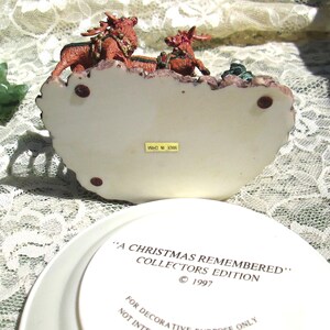 Może przedstawiać: Biała ceramiczna podstawa z figurką renifera w kolorze czerwonym i zielonym na g&oacute;rze. Podstawa zawiera napis "A Christmas Remembered" Collectors Edition &copy; 1997. Tylko do cel&oacute;w dekoracyjnych.