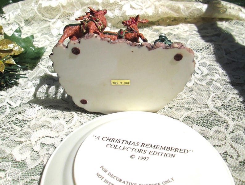 Może przedstawiać: Biała ceramiczna podstawa z figurką renifera w kolorze czerwonym i zielonym na g&oacute;rze. Podstawa zawiera napis "A Christmas Remembered" Collectors Edition &copy; 1997. Tylko do cel&oacute;w dekoracyjnych.