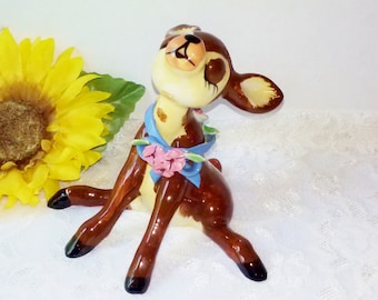 May include: A ceramic figurine of a brown and white deer with a blue ribbon and pink flowers around its neck. The deer is sitting up with its head tilted back and its eyes closed.