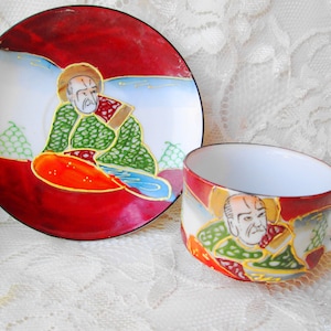 May include: A red and white teacup and saucer set with a gold trim. The set features a hand-painted design of a man in a green robe with a red and gold background.