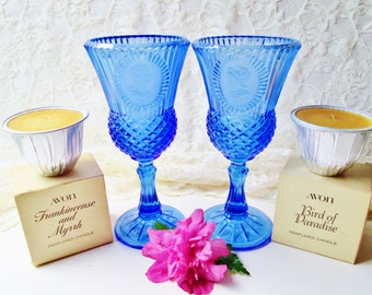 May include: Two blue glass goblets with diamond-cut detailing and a circular embossed design. Two small candles in foil containers and two boxes of Avon scented candles are also present. A pink flower is at the bottom.