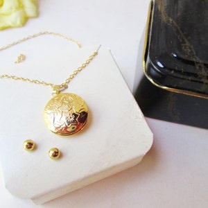 May include: A gold necklace with a round locket pendant featuring a floral design. The locket is open and the necklace comes with a pair of gold stud earrings.