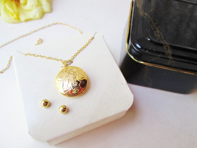 May include: A gold necklace with a round locket pendant featuring a floral design. The locket is open and the necklace comes with a pair of gold stud earrings.