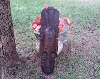 May include: A dark brown wooden mask with intricate carvings. The mask has a long, narrow face with a prominent nose and large eyes. The mask is decorated with a series of vertical lines and a feather-like design at the top.