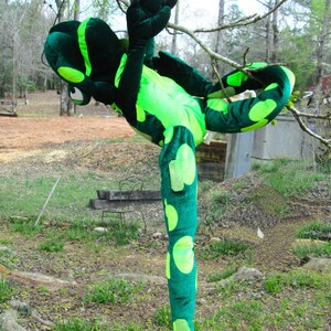 Pu&ograve; includere: Un costume da lucertola di peluche verde e giallo con una lunga coda e gambe macchiate. Il costume &egrave; in posizione eretta con una gamba sollevata.