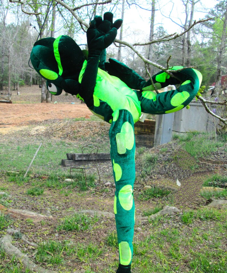 Pu&ograve; includere: Un costume da lucertola di peluche verde e giallo con una lunga coda e gambe macchiate. Il costume &egrave; in posizione eretta con una gamba sollevata.