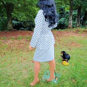 K&ouml;nnte beinhalten: Eine Frau in einem wei&szlig;en und schwarzen Polka-Dot-Shirt und einem grauen Rock steht auf einem gr&uuml;nen Rasen. Ein kleiner schwarz-brauner Hund sitzt auf dem Rasen mit einer gelben Gummiente.