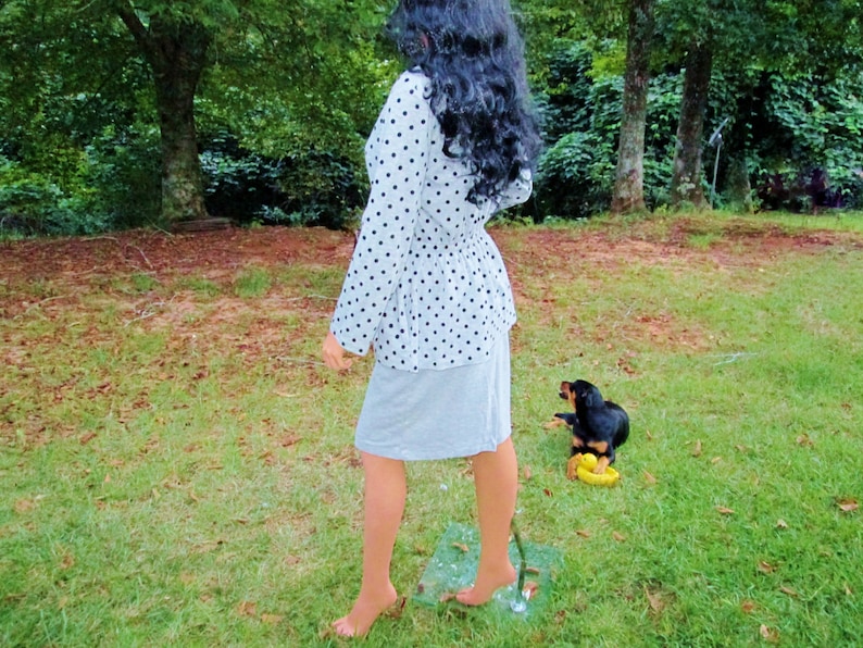 K&ouml;nnte beinhalten: Eine Frau in einem wei&szlig;en und schwarzen Polka-Dot-Shirt und einem grauen Rock steht auf einem gr&uuml;nen Rasen. Ein kleiner schwarz-brauner Hund sitzt auf dem Rasen mit einer gelben Gummiente.