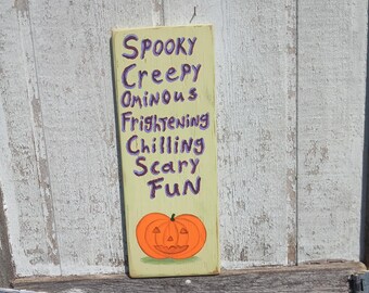 Op de afbeelding: Een rechthoekig, lichtgroen houten bord met de woorden "Spooky, Creepy, Ominous, Frightening, Chilling, Scary, Fun" in paars. Onderaan staat een oranje pompoen. Het bord staat op een verweerde, witte houten ondergrond.