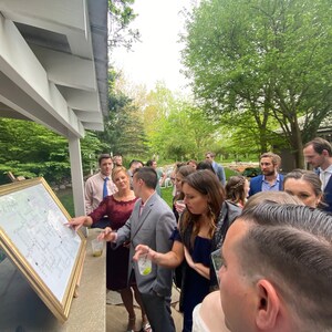May include: A group of people gathered around a large, framed, white board with gold trim. The board is covered in writing and appears to be a seating chart for a wedding. The people are looking at the board and talking to each other.