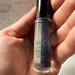 May include: A clear glass roller bottle with a black cap. The bottle has a label with a galaxy-themed design and the text "Love & Light" on it.