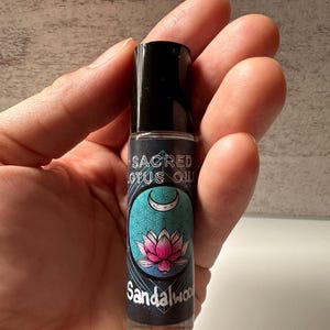 May include: A small glass bottle with a black rollerball top. The label is teal blue with a pink lotus flower and a crescent moon. The text on the label reads "Sacred Lotus Oil Sandalwood".