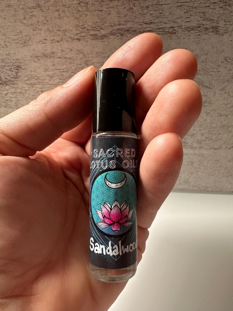 May include: A small glass bottle with a black rollerball top. The label is teal blue with a pink lotus flower and a crescent moon. The text on the label reads "Sacred Lotus Oil Sandalwood".