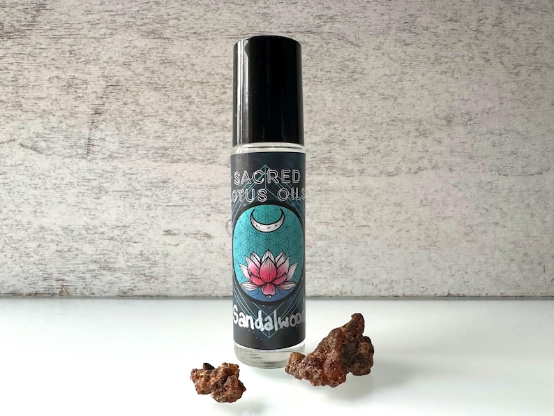 May include: A clear glass roll-on bottle with a black cap and a label that says "Sacred Lotus Oils" and "Sandalwood". The label features a pink lotus flower and a crescent moon on a blue and teal background. Two pieces of brown resin are in the foreground.