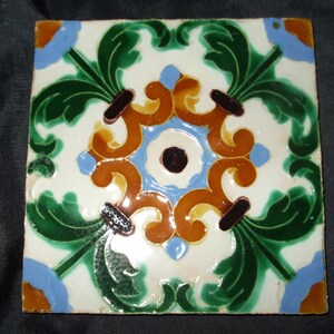 May include: A square ceramic tile with a decorative design featuring green, blue, brown, and yellow floral motifs. The tile has a white background and a glossy finish.