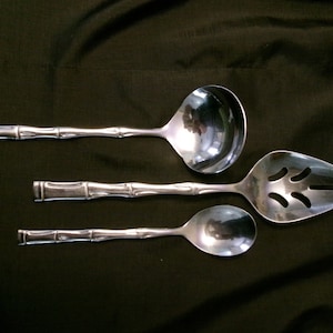 May include: Three silver kitchen utensils with bamboo-style handles. The utensils include a slotted spoon, a ladle, and a serving spoon.