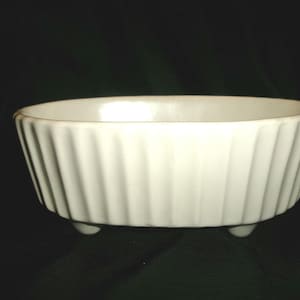 May include: White ceramic planter with a ribbed design and three small feet. The planter is oval shaped.