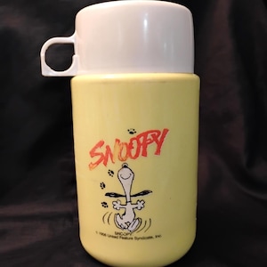 May include: A vintage yellow Snoopy thermos with a white lid and handle. The thermos features a red "Snoopy" logo and a graphic of Snoopy dancing. The bottom of the thermos reads "Snoopy © 1956 United Feature Syndicate, Inc."