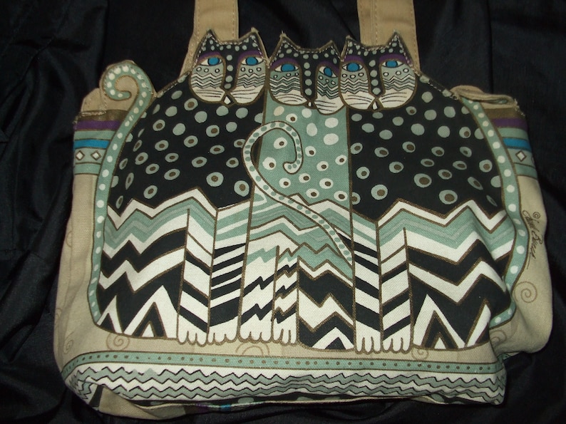 May include: A beige tote bag with a colorful design of three stylized cats. The cats are black, white, and green with gold accents. The bag has a geometric pattern and a whimsical design.
