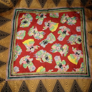 May include: A red silk scarf with a repeating pattern of colorful figures in traditional Chinese clothing. The scarf has a black, gold, and blue border.