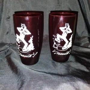 May include: Two deep red, translucent drinking glasses featuring a white graphic print of a person dancing with a dog. Below the figures, the text "Partners All" is printed in white, accompanied by small musical note symbols on both glasses.