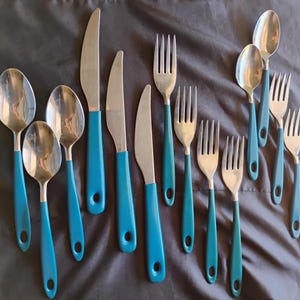 May include: A collection of stainless steel flatware with teal blue handles. The set includes spoons, forks, and knives. The handles have a rounded shape with a hole at the end. The flatware is arranged on a dark gray surface.