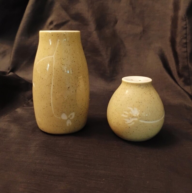 May include: Two ceramic vases. The taller vase is a light yellow with a speckled finish and white floral design. The shorter vase is also light yellow with a speckled finish and a white floral design. Both vases are on a brown background.