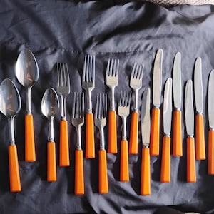 May include: A collection of stainless steel silverware with orange handles. The set includes spoons, forks, and knives, arranged on a dark gray surface. The silverware has a classic design, with a bright, solid orange handle.