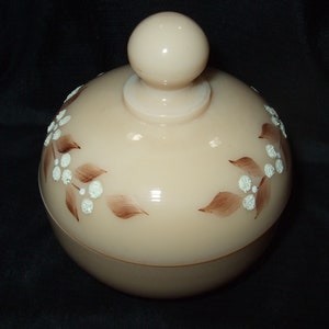 May include: A vintage glass jar with a lid, featuring a floral design in brown and white. The jar is a pale pink color and has a smooth, rounded shape.