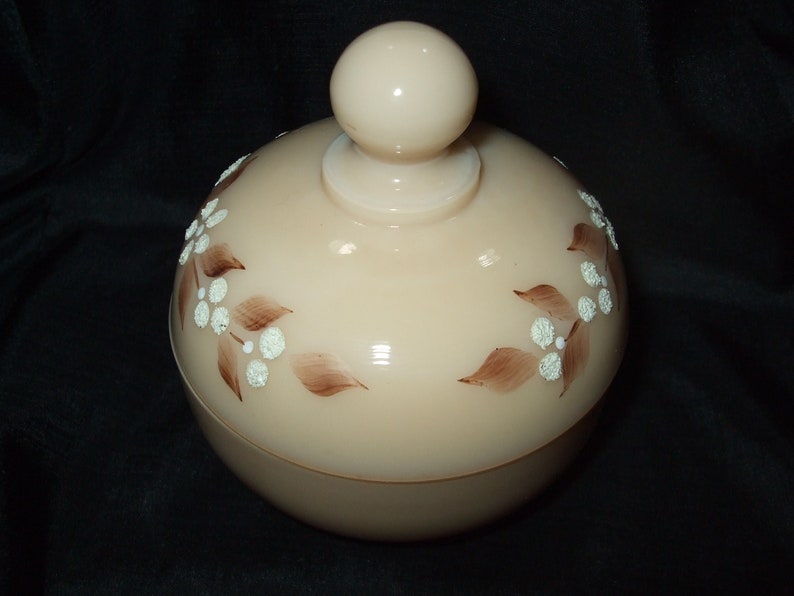 May include: A vintage glass jar with a lid, featuring a floral design in brown and white. The jar is a pale pink color and has a smooth, rounded shape.