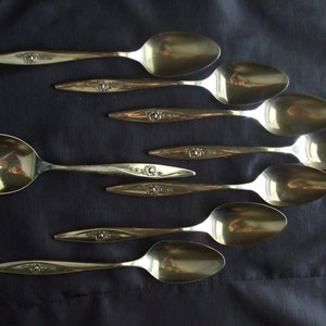 May include: A set of eight silver spoons with floral designs on the handles. The spoons are arranged in a row, with the largest spoon at the top and the smallest spoon at the bottom.