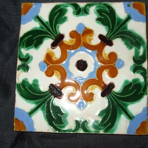 May include: A square ceramic tile with a decorative design featuring green leaves, blue and orange swirls, and a black and white center.