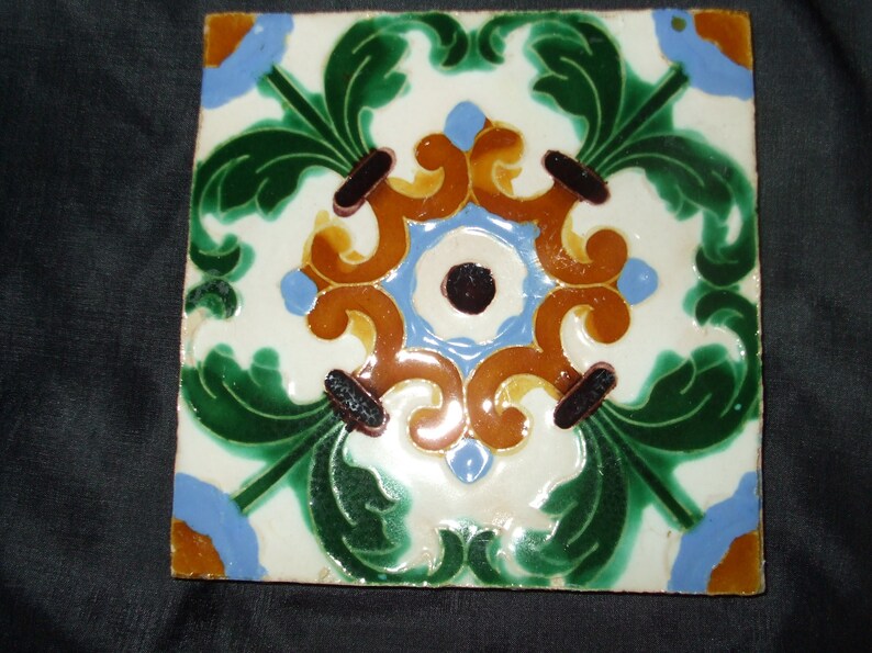 May include: A square ceramic tile with a decorative design featuring green leaves, blue and orange swirls, and a black and white center.