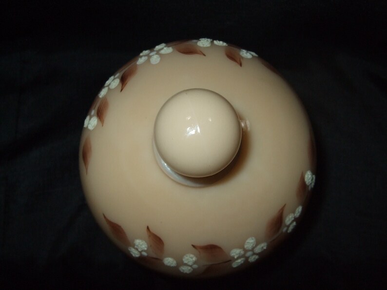 May include: A pale pink glass jar with a white knob lid. The jar is decorated with a floral pattern of white flowers and brown leaves.