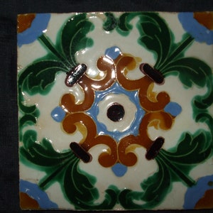 May include: A square ceramic tile with a white background and a colorful floral design. The design features green leaves, blue and brown swirls, and a black and white center.